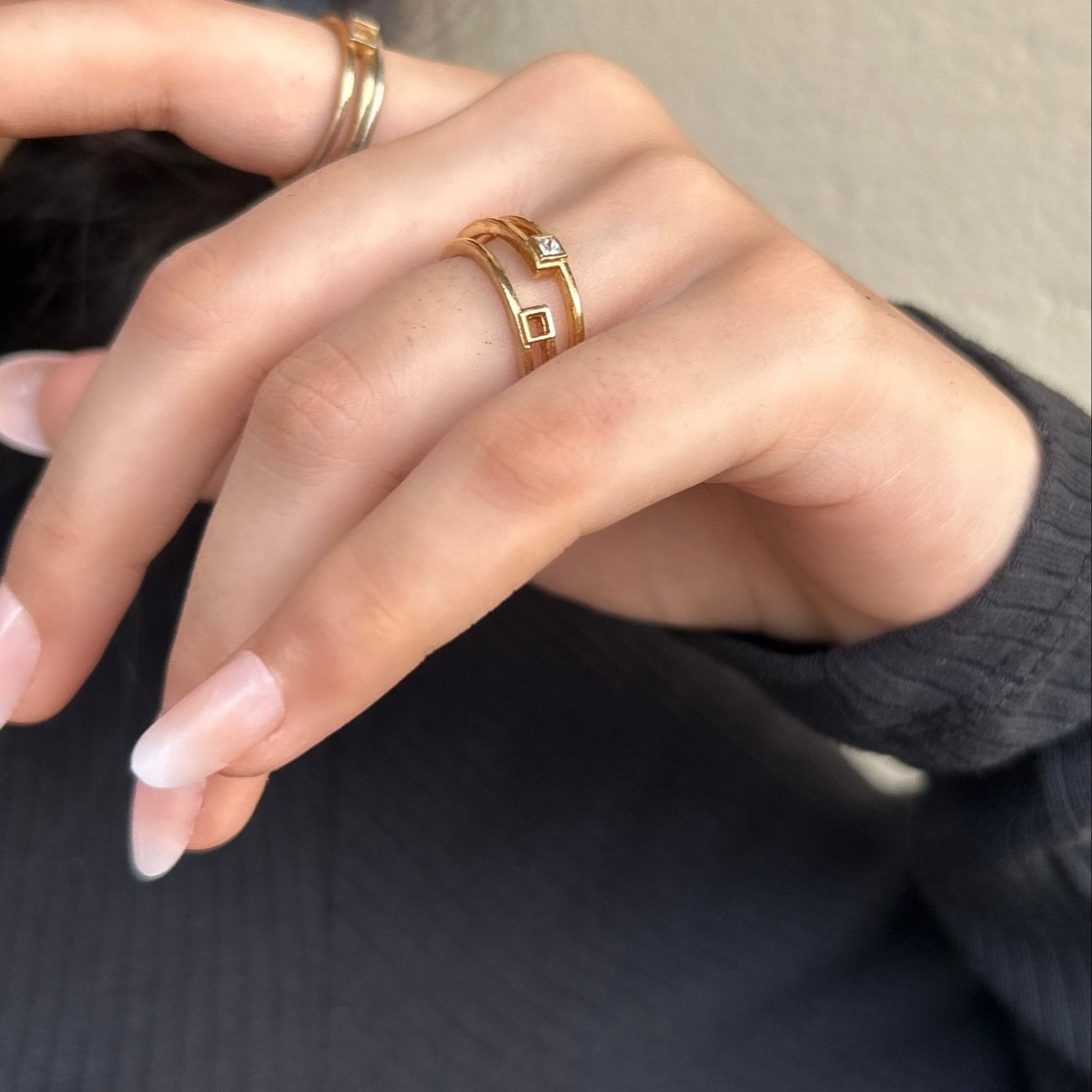 Close-up of a hand with gold rings and pink nail polish, wearing a dark gray sweater.