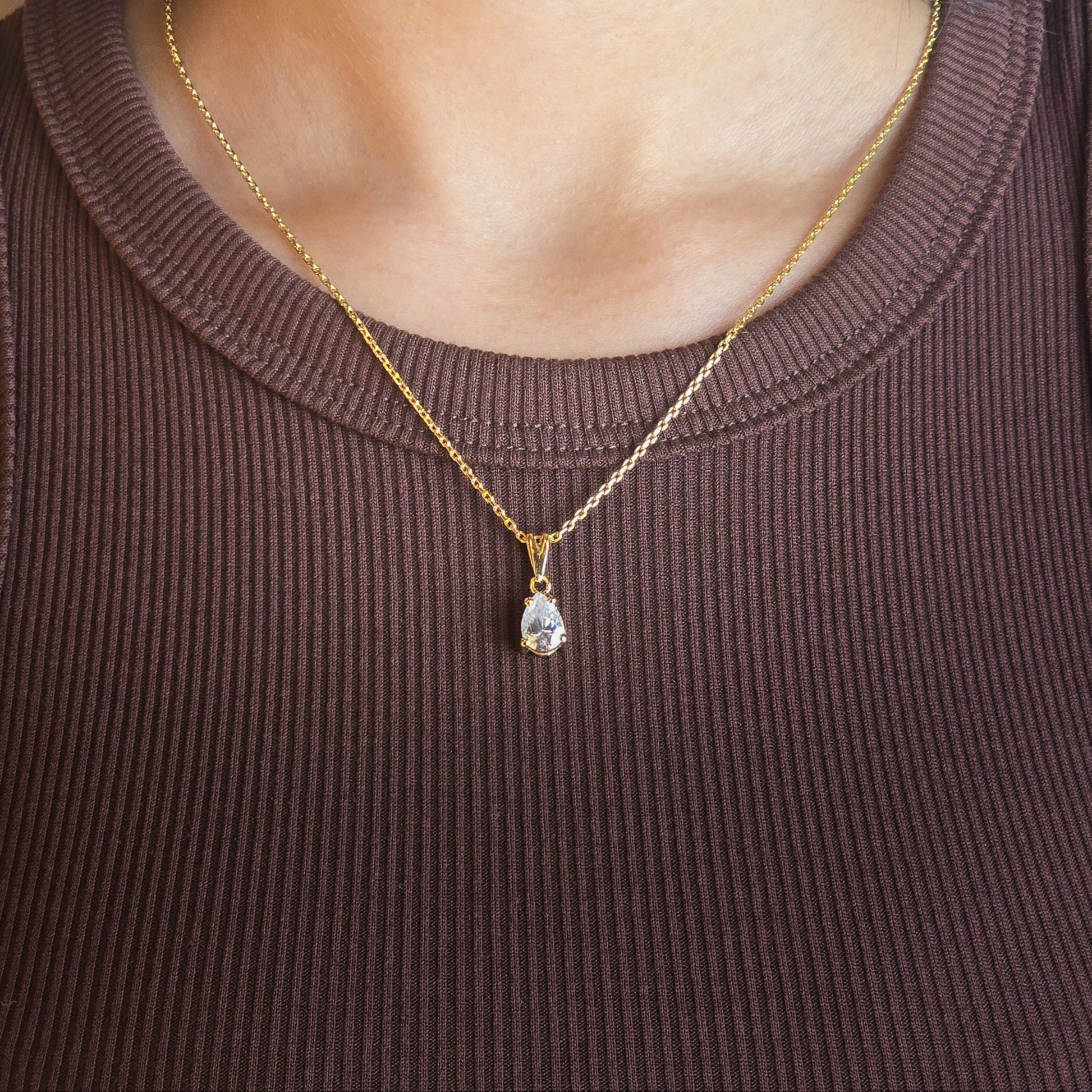 Person wearing a gold necklace with a pendant on a brown sleeveless top.
